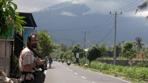 Mark Levitin, 39, a freelance photographer from Russia, stands on a street with a backdrop of the Mount Agung volcano covered by cloud in Karangasem, Bali. (AAP)
