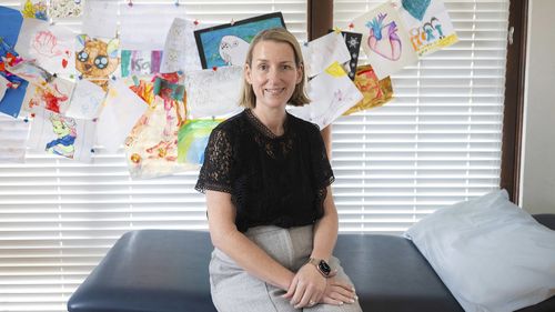 La dottoressa Rebekah Hoffman siede su un lettino nel suo studio di Sydney. Indossa pantaloni grigi, una camicia nera e sul muro dietro di lei ci sono disegni di bambini.