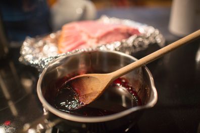 Close up photo of a pot with a wine reduction glaze in it. The silver pan has a wooden spoon and you can see the deep red sauce on it. There are rosemary leaves in it. In the background you can see ham laying on tin foil, but it is out of focus. the shot has a very narrow depth of field. 