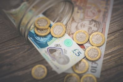 A stock photo of some British Money spilling out of a glass jar on a wooden background. Showing a Five and Ten pound note with the new One Pound coins.