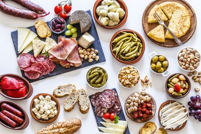 Large variety of spanish tapas shot from above on white background. The composition includes spanish tortilla, pickles, cheese, chorizo, bread, peanuts, pistachios, salami, prosciutto, jalapeño peppers, anchovies and others. 