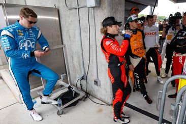 Josef Newgarden stands alone, looking at his phone, while other IndyCar drivers talk together.