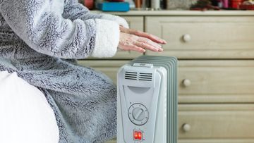 A retired senior woman in her 70s sits at home inside her cold house in winter. It is so cold that she is wrapped up in warm winter clothing, and is holding her hands over an electric heater for some extra warmth and comfort. Selective focus with room for copy space.
