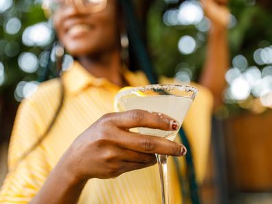 Low angle view of cheerful young woman holding and enjoying a martini glass with margarita cocktail during a relaxing summer garden party. Part of a series.