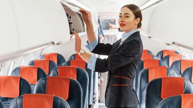 Young stewardess that is in formal black clothes is standing indoors in the plane.