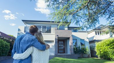 Couple standing in front of their new home. They are both wearing casual clothes and embracing. Rear view from behind them. The house is contemporary with a brick facade, driveway, balcony and a green lawn. The front door is also visible. Copy space