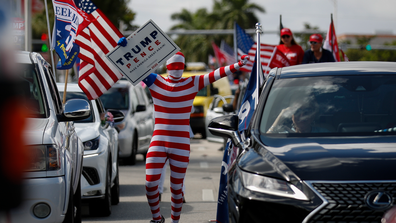 A man wearing a flag-themed body stocking walks between lines of cars as hundreds of vehicles gather for a car caravan in support of President Donald Trump, at Tropical Park in Miami, Sunday, Nov. 1, 2020. (AP Photo/Rebecca Blackwell)