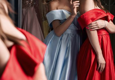 Two young beautiful women wearing off-the-shoulder full-length sky blue and crimson red satin slit prom ball gowns. Models looking in mirror.