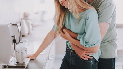 Bearded guy standing behind blonde girl and kissing her. They are waiting for hot drink