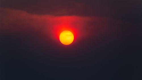 The ghostly and obscured sun shrouded in dense, orange-grey smoke from a devastating forest fire. The summer sun struggles to penetrate the haze, casting an eerie glow.