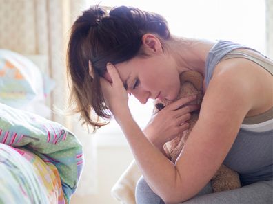 A grieving mother sits in her child's bedroom holding their toy in her arms while resting her head in her hands. The child's bed can be seen in the background untouched from when they were last in it.