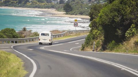 A Volkswagen Kombi (or Transporter) van driving down Great Ocean Road, with Lorne Beach in the background.