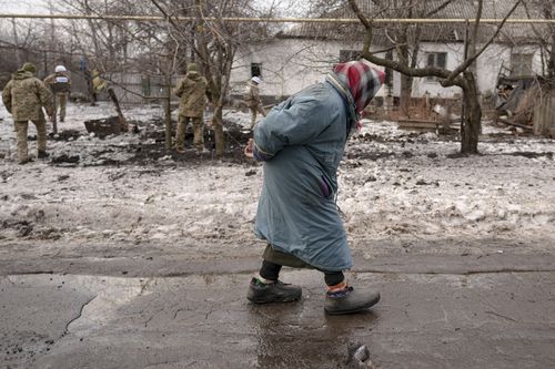 An elderly lady walks by as members of the Joint Centre for Control and Coordination on ceasefire of the demarcation line, or JCCC, survey a crater and damage to a house from artillery shell that landed in Vrubivka, one of the at least eight that hit the village today, according to local officials, in the Luhansk region, eastern Ukraine, Thursday, Feb. 17, 2022.  