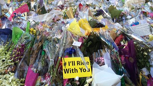 Cyclists lay flowers at Sydney siege memorial with #I'll ride with you signs.