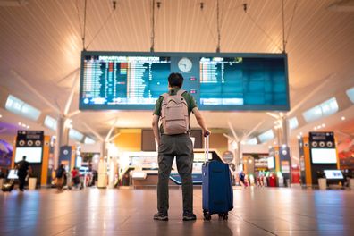 Asian male traveler with wheeled luggage checking for flight schedule on arrival and departure board at Kuala Lumpur International Airport