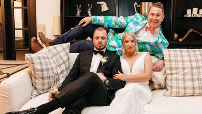 The Colourful Celebrant Brad with his two clients, dressed and ready for their wedding. The couple sit on a couch facing the camera bare faced, while Brad lays behind them in a bright aqua suit jacket with pink flamingos printed on it.