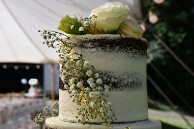 White gypsophila (or baby's breath) and white roses decorate the top tier of a white and chocolate wedding cake. A cream macaroon with yellow filling is among the fowers. The focus is on the foreground, with a table and the pavilllion out of focus in the background.