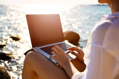 Modern young Caucasian female freelancer, working on laptop, from the rocky beach, during her vacation