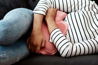 Cropped shot of a woman holding a hot water bottle against her stomach on the sofa at home