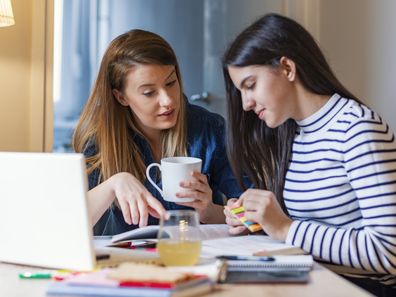 Photo of young brown hair Caucasian mother is helping her teen  daughter with her homework in front of laptop. They are looking away from the camera. Dark hair schoolgirl and mother together doing homework in home. Beautiful Hispanic girl and her young mother reading a book together or studying at home during the day.