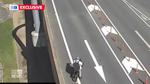 In CCTV vision, a cyclist is captured heading towards oncoming traffic before crossing over two lanes near a busy motorway tunnel.