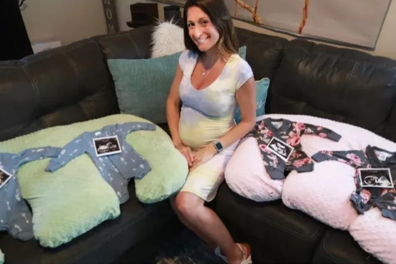 Ashley Ness sitting on the couch with four onesies laid out, two blue and two pink, with ultrasound pictures on top.