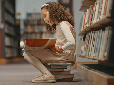 Young girl sitting on books in the library and reading a book. Cute girl with curly hair doing her project. Female alone and doing research for a project