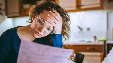 Woman sitting at kitchen table in front of open laptop, looking at a piece of paper in her hand and holding her head in her other hand