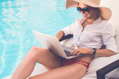 Smiling business woman in sunglasses and straw hat sitting by swimming pool and working on laptop