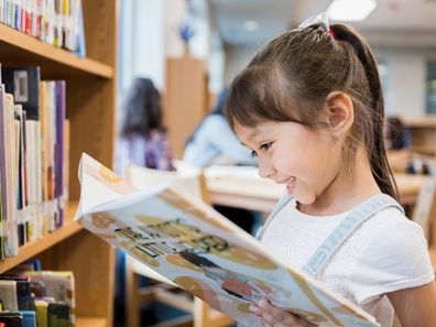 Adorable Hispanic schoolgirl smiles while delight as she reads a picture book while standing in the school library.