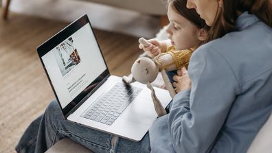 Working mum sits on her laptop with her child. Stock photo.