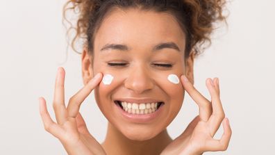 Skin care. Laughing girl applying moisturising cream on her face over white background