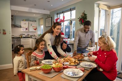 A front view of a family getting settled to have Christmas dinner. They are setting the table with the food and all of the vegetables. There are also Christmas crackers on the table and the youngest little girl looks full of anticipation and excitement at the food.