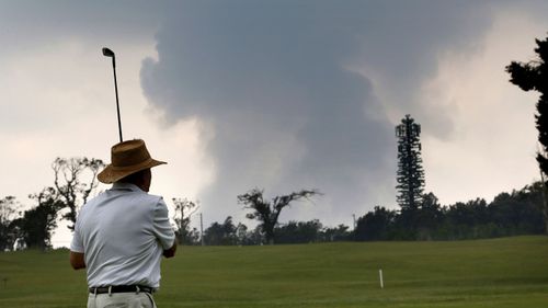 A man plays golf as as ash from the summit crater of Kilauea volcano rises in the background.