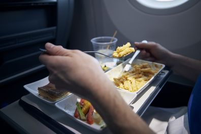 Passenger eating airline meal with metal cutlery. Menu with pasta, vegetable, dessert and drink on tray during long haul flight.