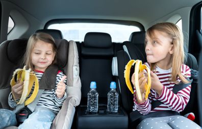 Little girls, sisters are driving in car, eating banana. Traveling on road in safe baby seats with child belts. Fun family trip, activity with parents.
