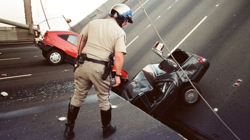 În această fotografie de fișier din 1989, un ofițer din California Highway Patrol inspectează daunele aduse mașinilor care au căzut atunci când puntea superioară a podului Bay s-a prăbușit pe puntea inferioară după cutremurul Loma Prieta din San Francisco. 