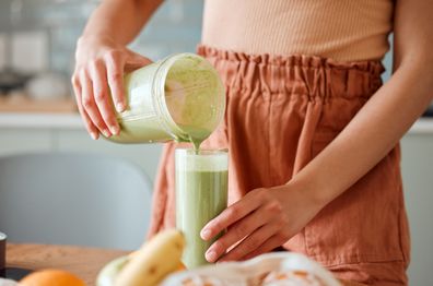 Woman pouring healthy smoothie in a glass from a blender jar on a counter for detox. Female making fresh green fruit  juice in her kitchen with vegetables and consumables for a fit lifestyle.  stock image