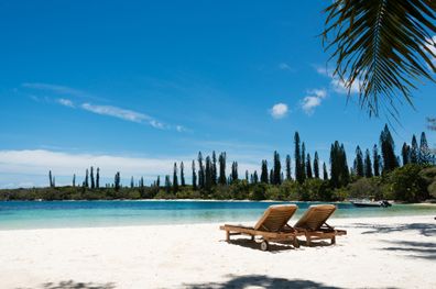 Kanumera beach on the Isle of Pines in New Caledonia. There are a pair of beachchairs on the white sand. The bay is surrounded by pacific cedars and is known as one of the most beautiful bays in the world.