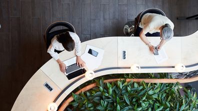 Young brunette woman and a senior lady using their devices while sharing a desk and keeping social distance office