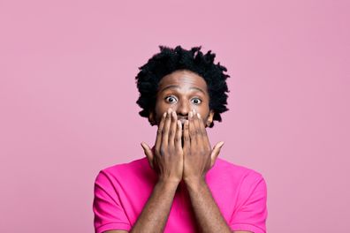 Summer portrait of surprised afro american young man wearing pink polo shirt, staring at camera and covering mouth with hands. Studio shot on pink background.