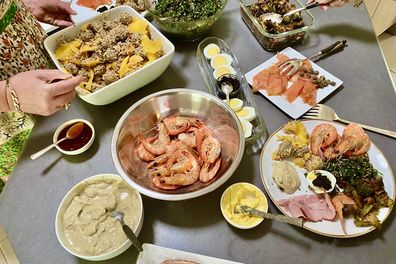 Horizontal spread of a variety of Christmas lunch delicacies on bench in domestic kitchen with hands serving and filling plates including fresh seafood of prawns oysters smoked salmon ham dips tabouli meatballs eggs and more