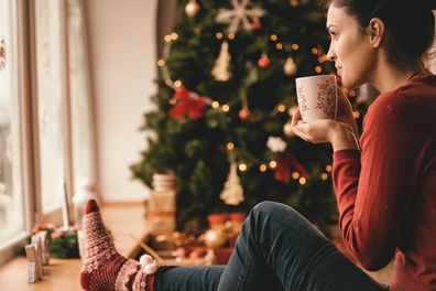 Young woman drinking tea by the Christmas tree, looking through window.