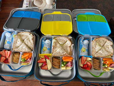 Three bento style school lunch boxes packed with sandwiches, fruit, vegetables and snacks