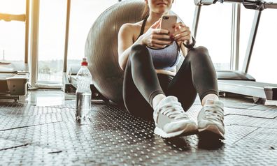 Young woman athlete using cell phone at gym. Young woman in sportswear checking phone while resting after workout on floor. Beautiful fit girl messaging with smartphone at fitness centre.