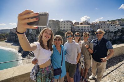 Multi generation family enjoying Calabrian town of Tropea on a sunny day. They are standing on the viewpoint by the Santuario di Santa Maria dell'Isola di Tropea and taking selfies. The beautiful cliff of Tropea is visible in the background. Shot with Canon R5