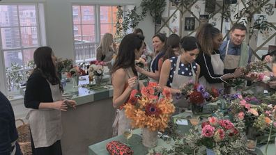 Meghan Markle, Amal Clooney, Serena Williams and others at bouquet arranging workshop at Markle's baby shower in 2019 in New York. Flowers range from a pastel orange, to deeper orange with some pink and purple flowers also present.