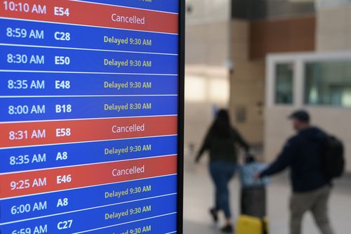 I viaggiatori camminano mentre una scheda video mostra i ritardi e le cancellazioni dei voli all'aeroporto nazionale Ronald Reagan di Washington ad Arlington, in Virginia, mercoledì 11 gennaio 2023.