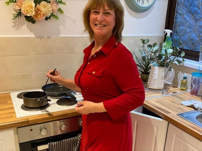 Older woman with shoulder length hair in red dress cooking on a stove and smiling at camera.