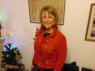 Older woman with shoulder length hair in red jacket smiling for the camera besides a Christmas tree.